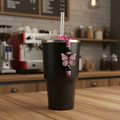 Pink and black butterfly beaded straw charm on a sleek matte black tumbler in a warm cozy coffee house setting